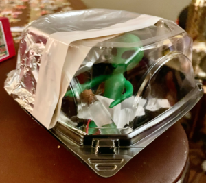 Model UFO made w/takeout container covered in foil. Green alien with large eyes is visible through plastic.